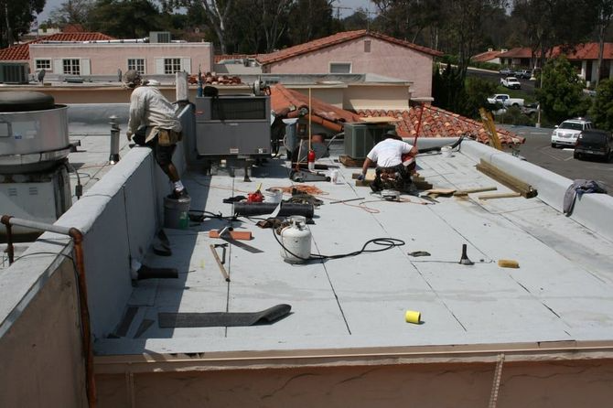 Two roofers work on a flat roof with tools and a propane tank under the sun.