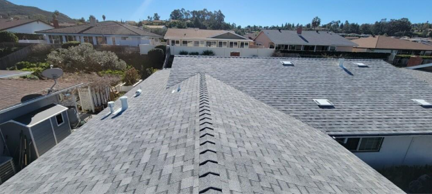 View of a gray shingled roof with a row of shingles running down the center, houses and blue sky in background.