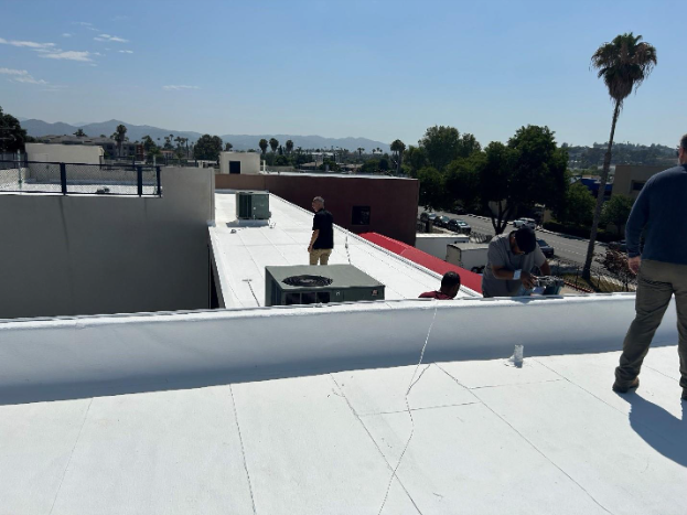 People working on a white flat roof with AC units, buildings, and a palm tree in the background.