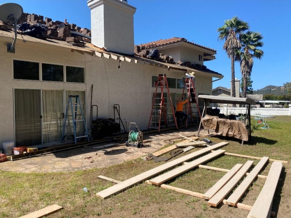 Workers on a roof removing shingles, tools and materials on the ground, sunny day.