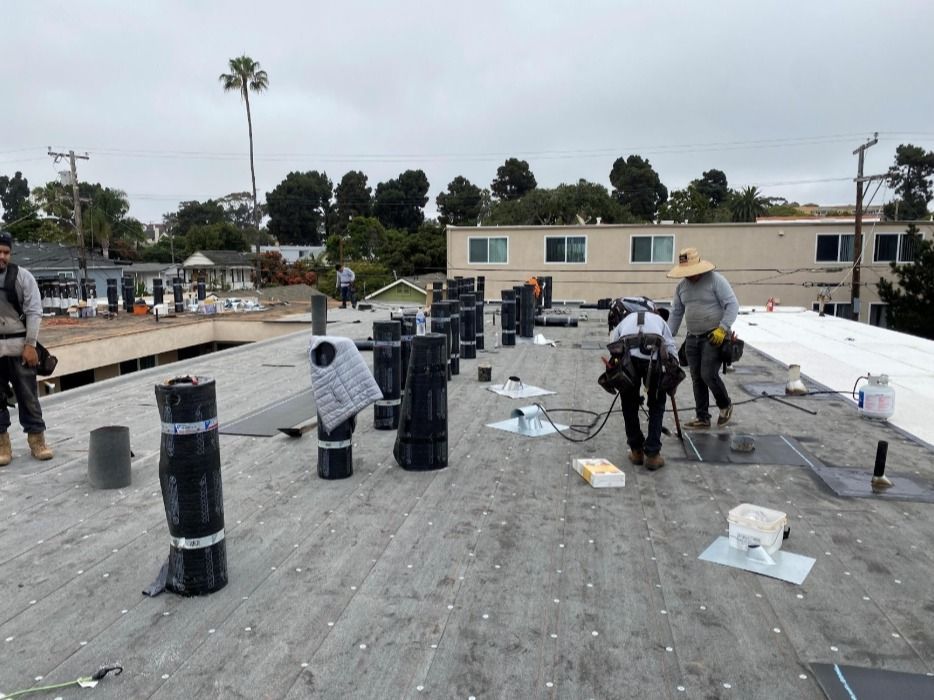 Roofers work on a flat roof; several upright rolls of roofing material stand alongside them, cloudy sky overhead.