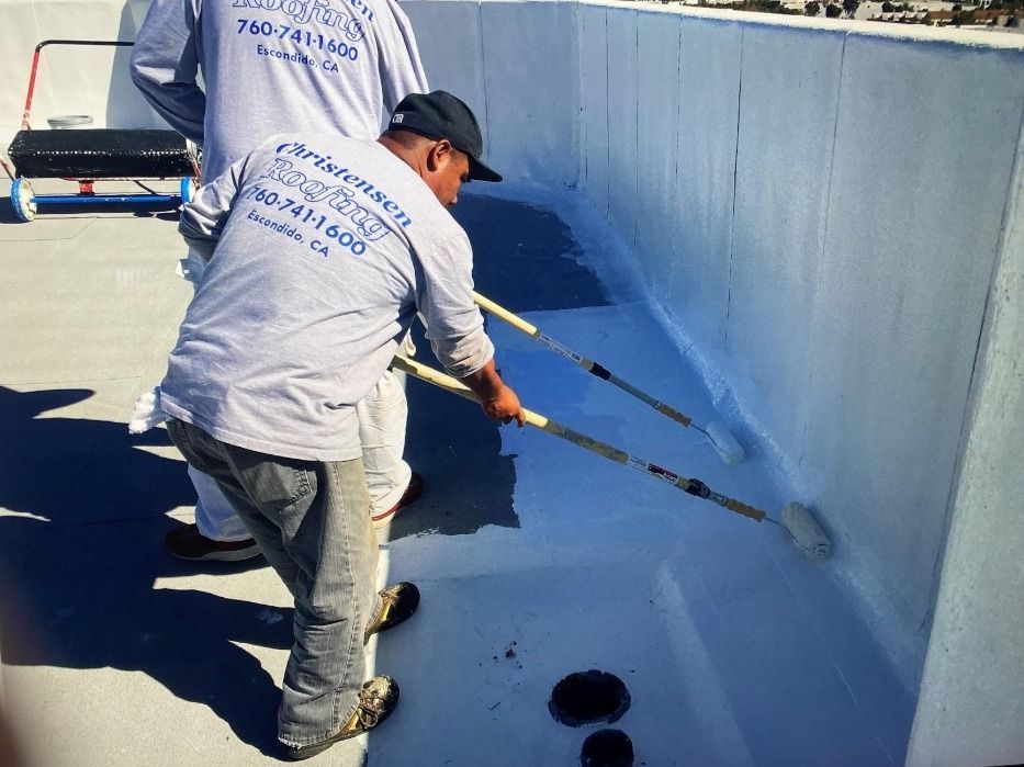 Two workers painting a light-colored roof with rollers; one wearing a cap, in Escondido, CA.