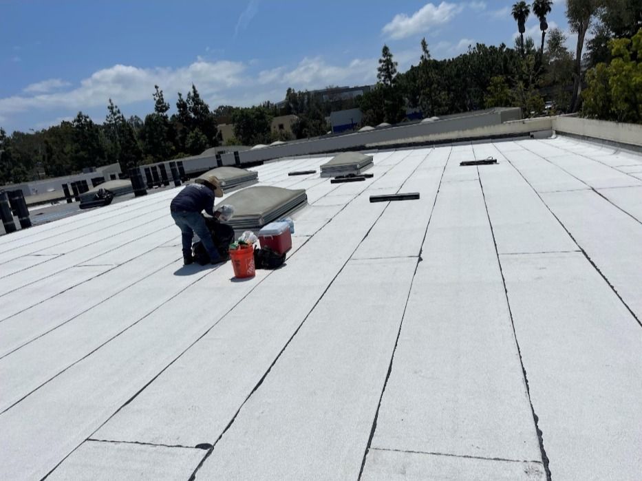 A person on a white flat roof, with equipment, possibly repairing or installing roofing material; outdoors, sunny.