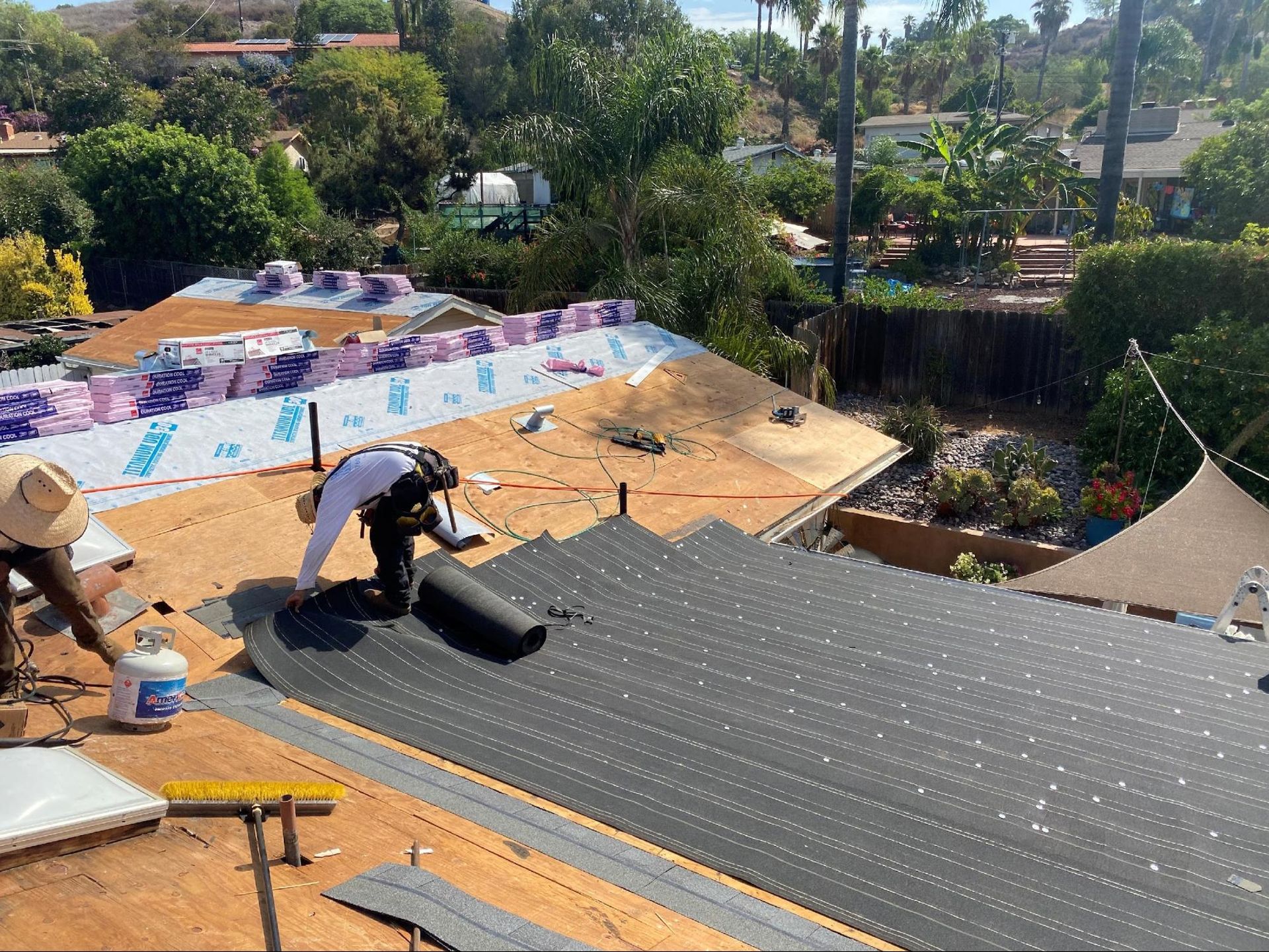 Two roofers installing a roof, one rolls out roofing material, the other works with a torch, outdoors, sunny day.