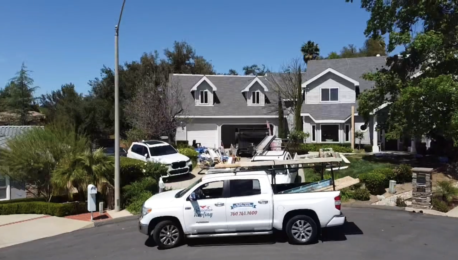 White truck parked in front of a gray house, work in progress. Blue sky, trees, and bushes.