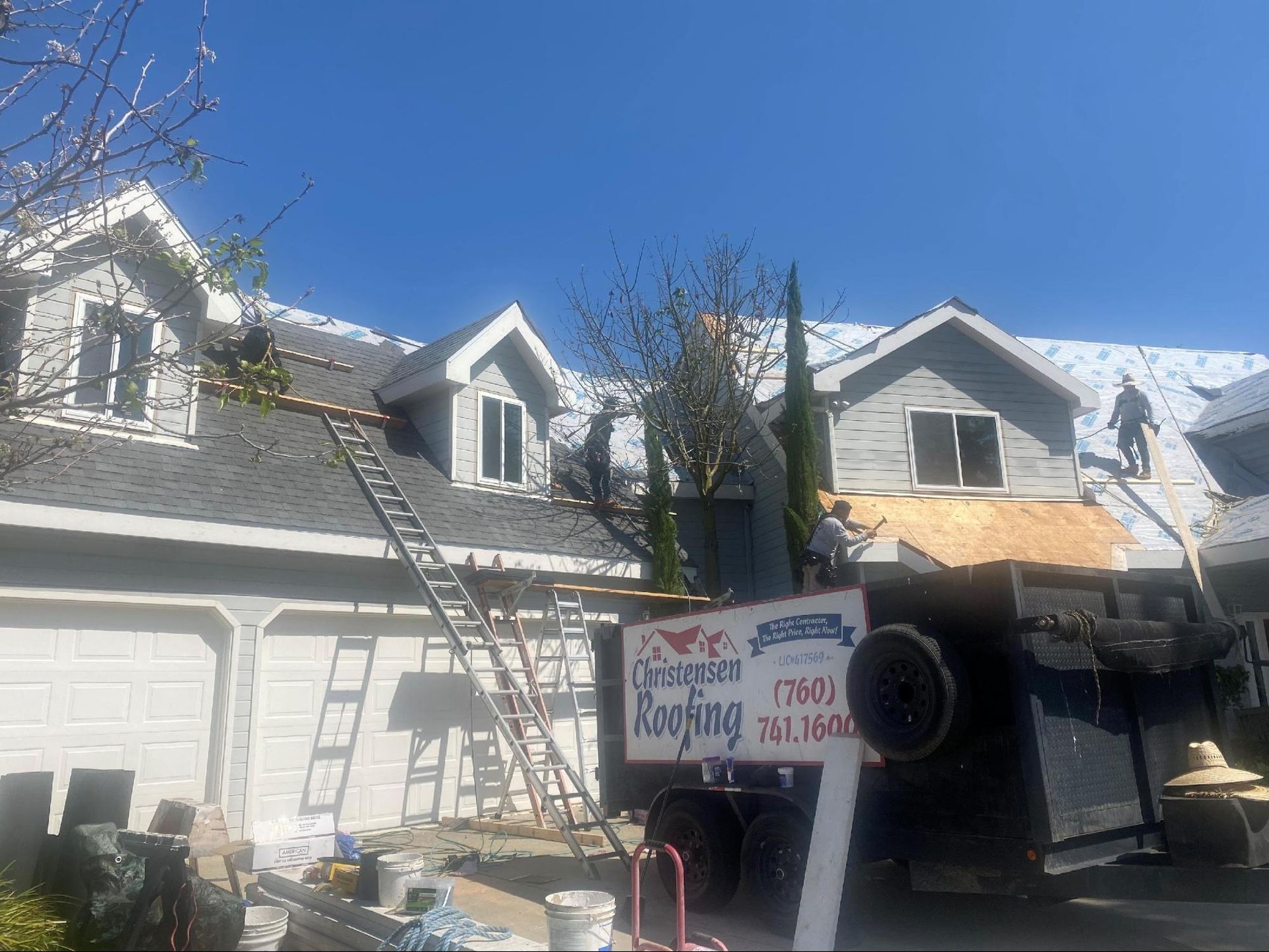 Roofers on a house with ladders and a dumpster, removing and replacing shingles. Clear blue sky.