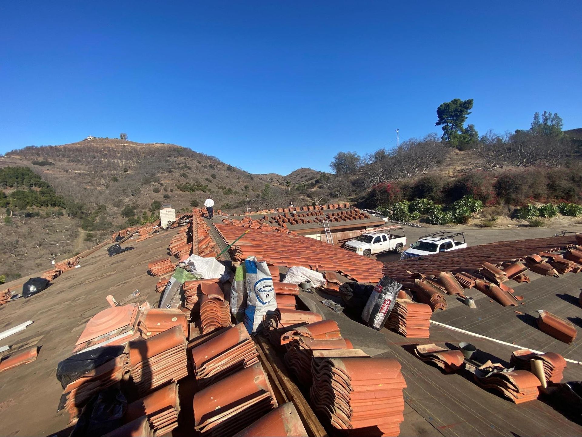 Roof with scattered red tiles, worker, vehicles, and hill under a clear blue sky.