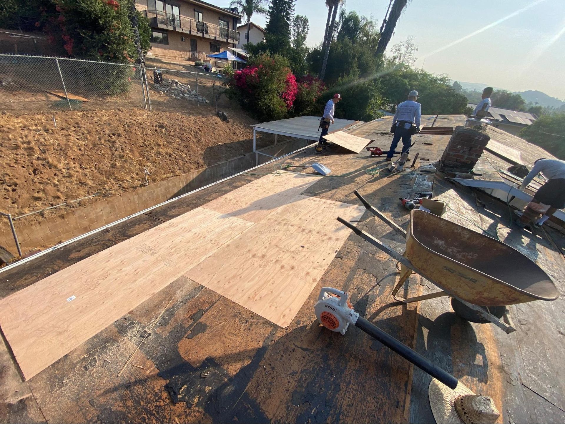 Workers installing plywood on a rooftop with a wheelbarrow and hillside in the background.