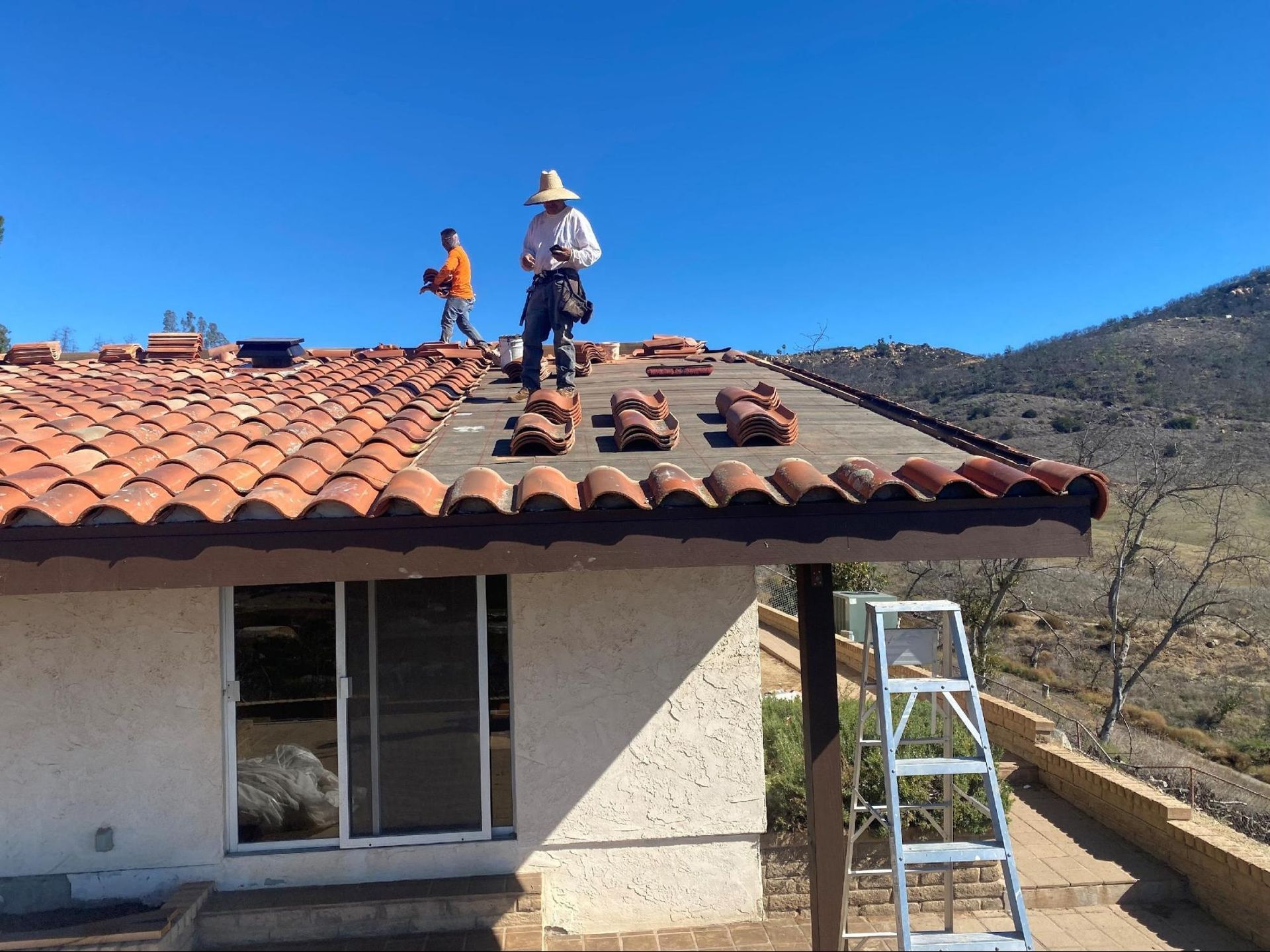 Two roofers on a terracotta tiled roof, removing tiles. Sunny day, mountains in background.