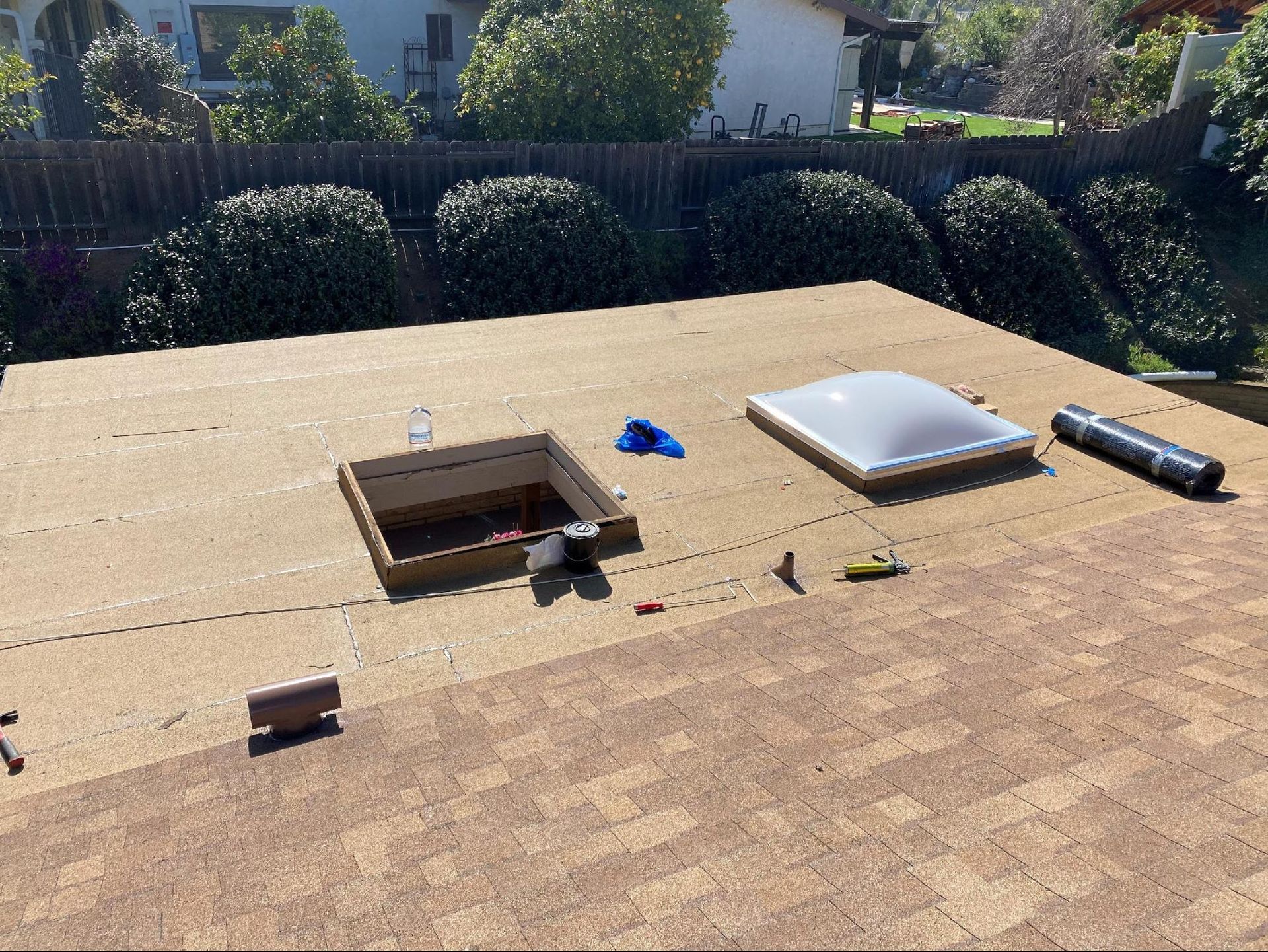 Flat rooftop with skylight and opened vent surrounded by shrubbery and fence.