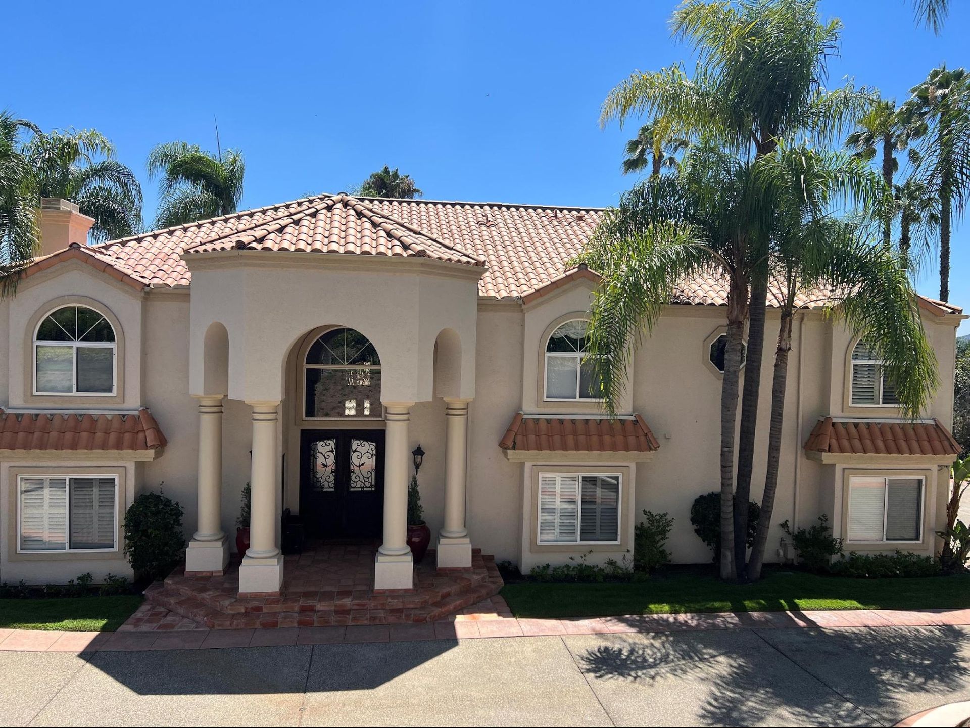 A large, beige stucco house with red-tiled roof, arched windows, and palm trees under a blue sky.