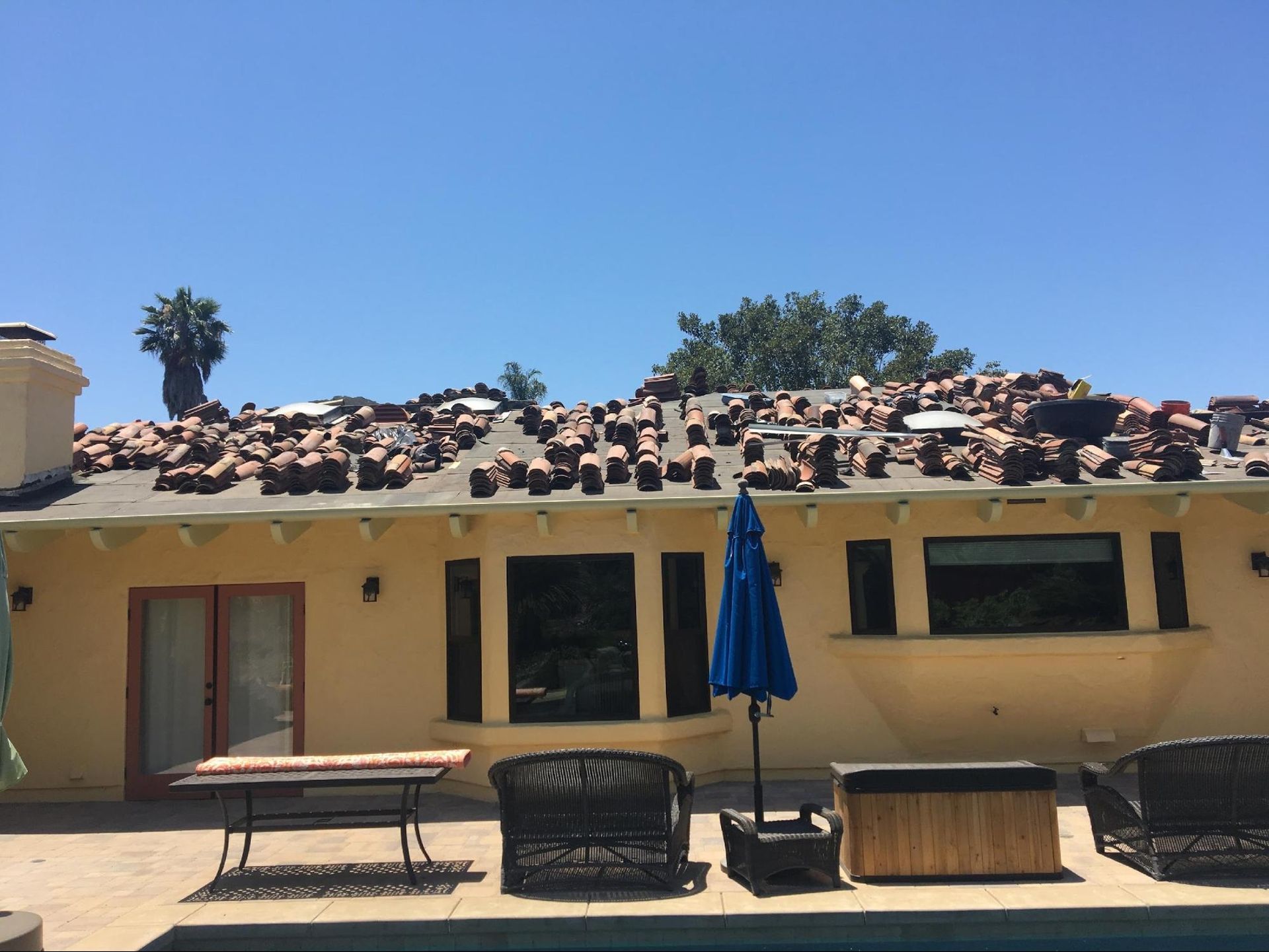 Yellow house with roof tiles scattered on the roof, blue umbrella, pool in front.