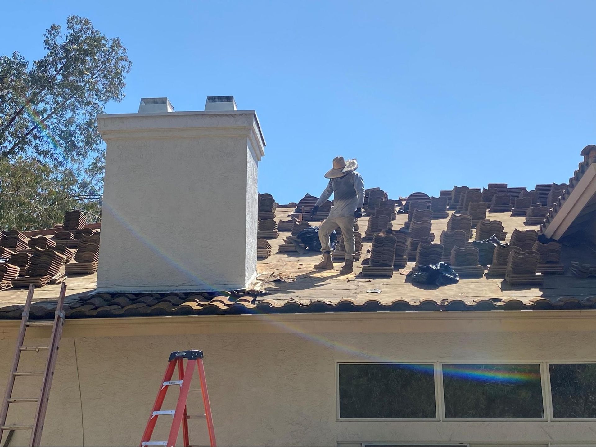 A worker on a roof removing clay tiles.  Ladder and chimney visible against a blue sky.