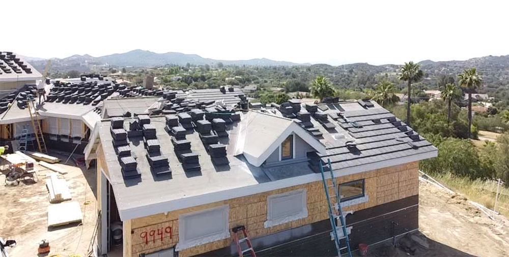 House under construction, roof partially shingled, surrounded by stacks of shingles and scenic background.