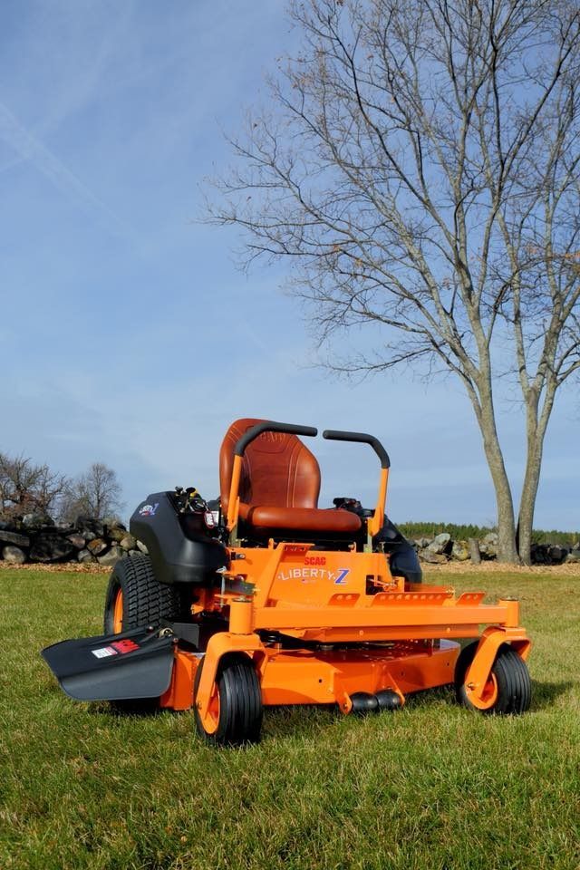 Orange zero-turn lawn mower on grassy field. A leafless tree stands in the background under a blue sky.