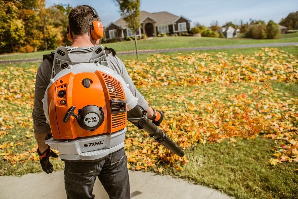 Man using a backpack leaf blower on a lawn with fallen leaves near a house.