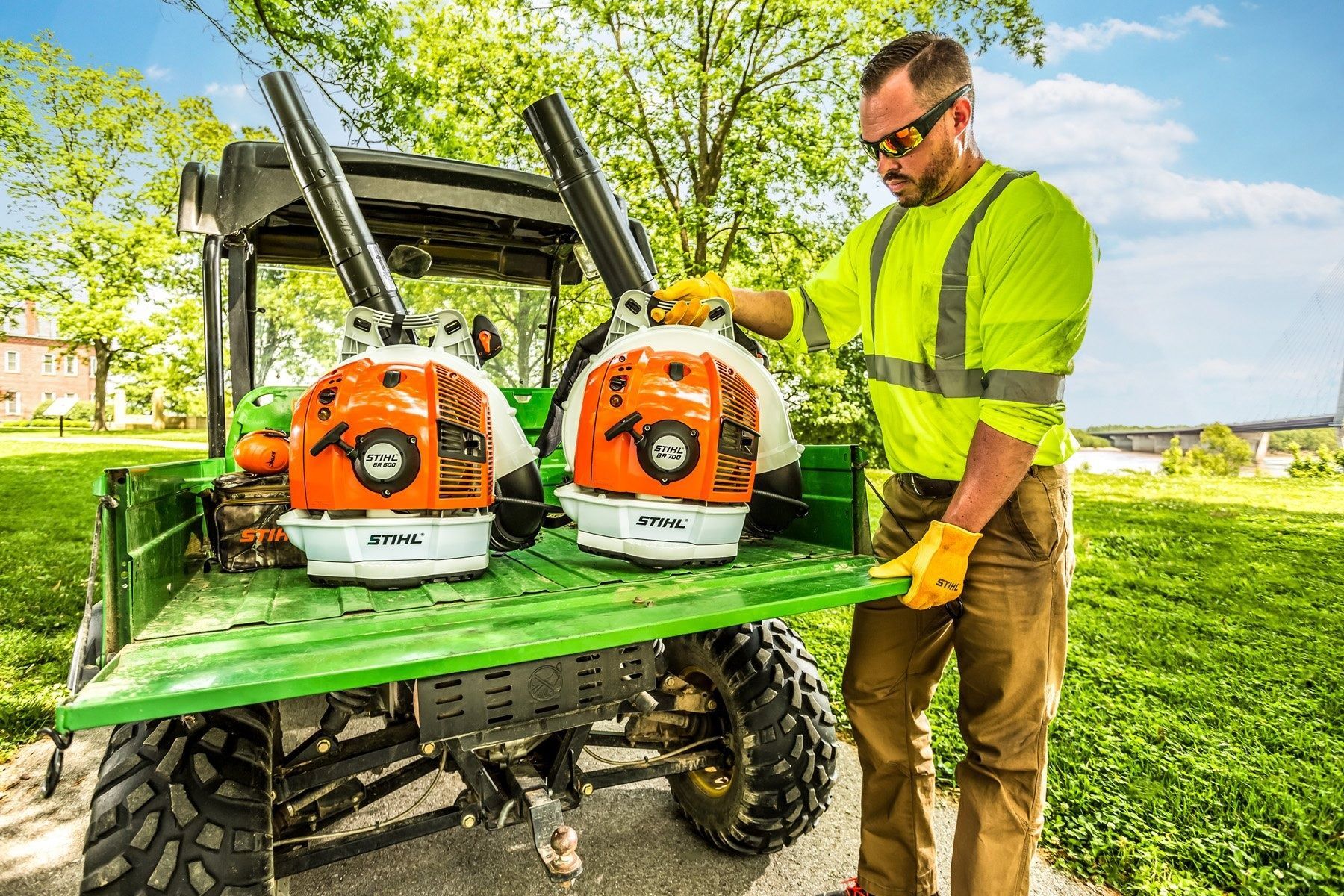 Man in safety vest loads two leaf blowers onto a utility vehicle in a park.