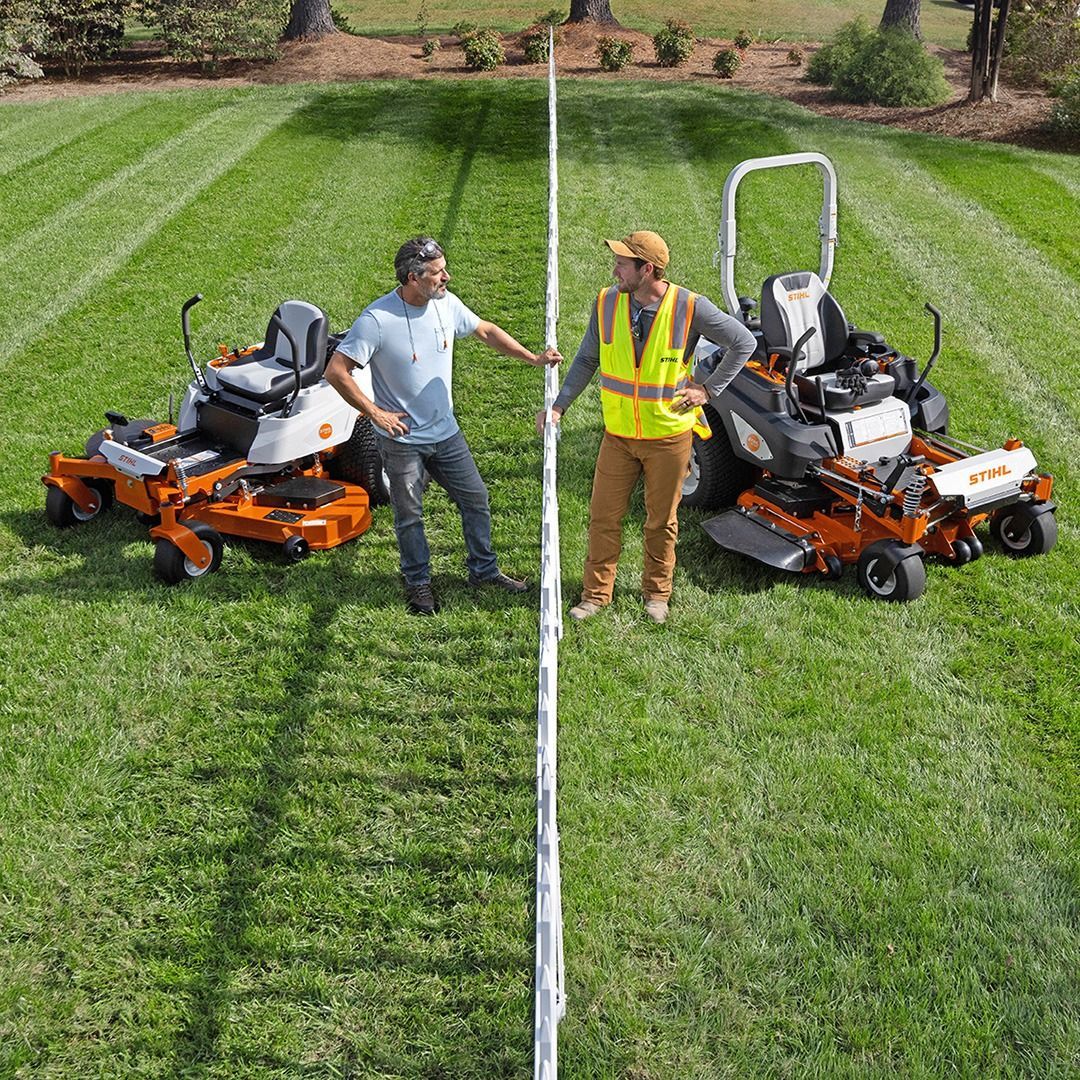 Two people beside riding lawnmowers measure straightness with a long pole in a green yard.