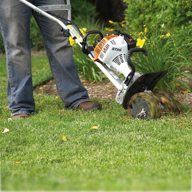 Person in jeans and work boots using a string trimmer to edge a flower bed.