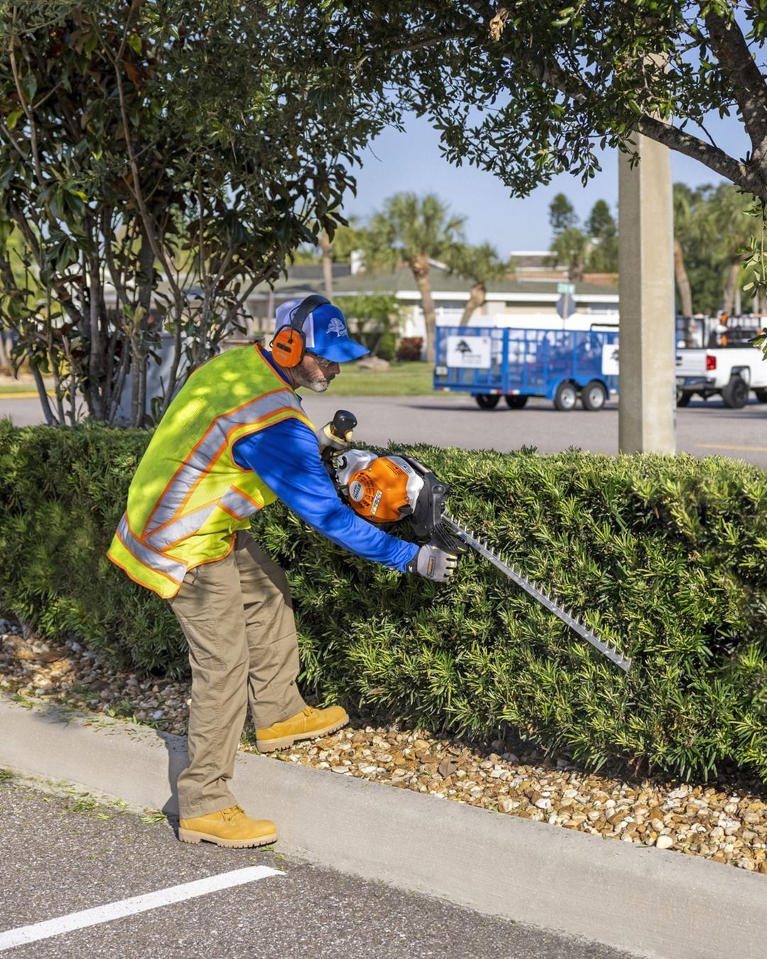 A person in safety gear trims a hedge with a power trimmer near a parking lot curb.