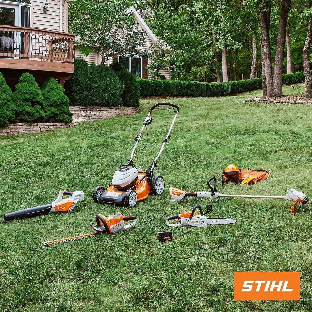 Lawn care equipment on a grassy lawn with a house in the background. Tools include a lawnmower, blower, chainsaw and trimmer.