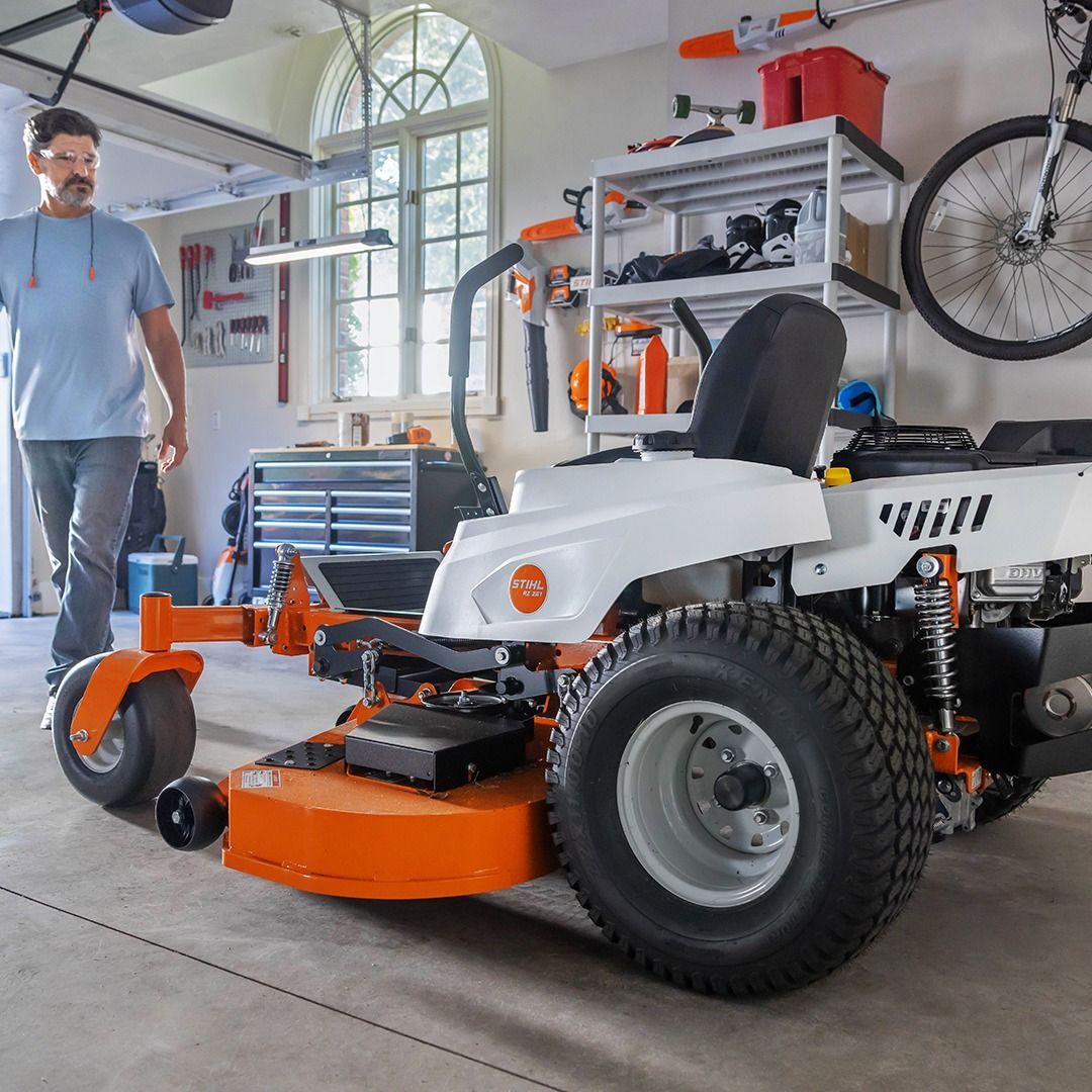 Man stands beside an orange and white riding lawnmower in a garage.