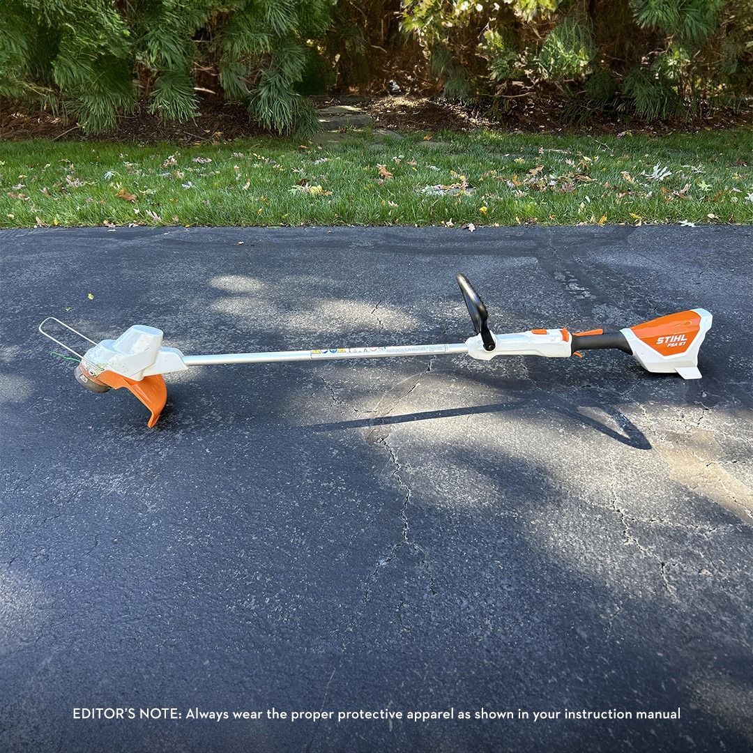 A Stihl string trimmer lies on asphalt in front of a green lawn and bushes.
