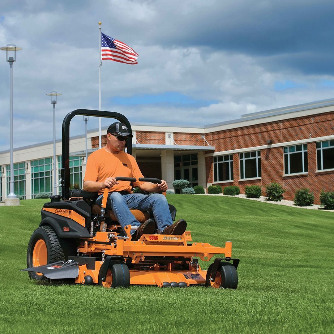 Man operating a bright orange zero-turn mower on a grassy lawn in front of a building with an American flag.