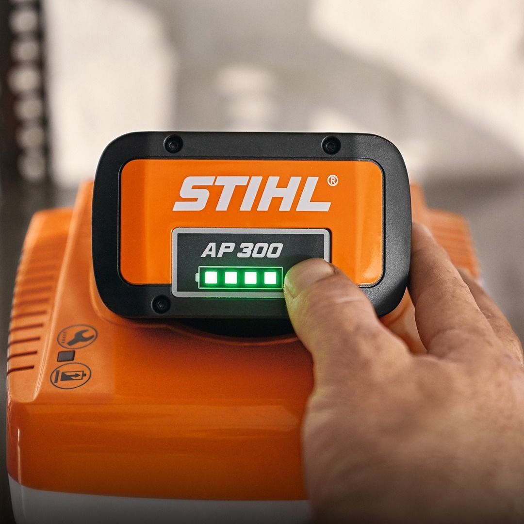 Hand pressing a button on a Stihl AP 300 battery displaying four green lights, on an orange charger.