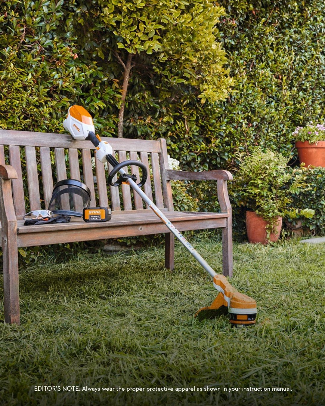 Lawn trimmer, battery, and safety gear on a wooden bench in a garden.