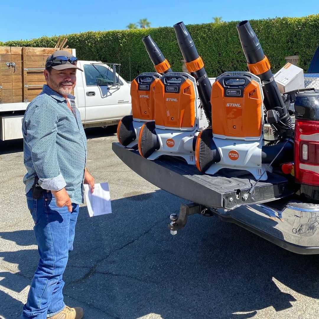 Man stands beside a truck bed with three orange and gray leaf blowers. He holds a paper and smiles.