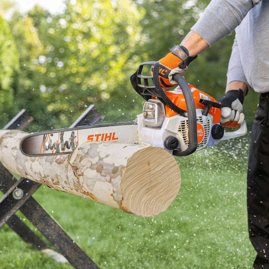Person using an orange and white chainsaw to cut a log on a sawhorse in a grassy outdoor setting.