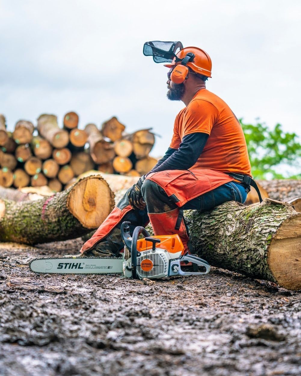 Lumberjack resting on a log with a chainsaw, wearing safety gear and looking away. Logs and cloudy sky.