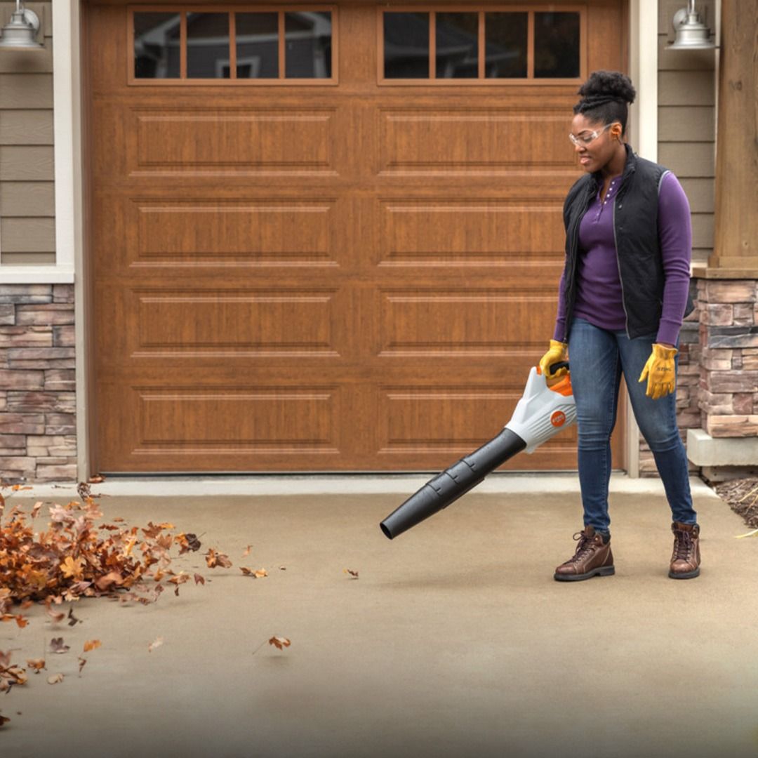 Woman using a leaf blower on a concrete driveway in front of a garage, blowing leaves.