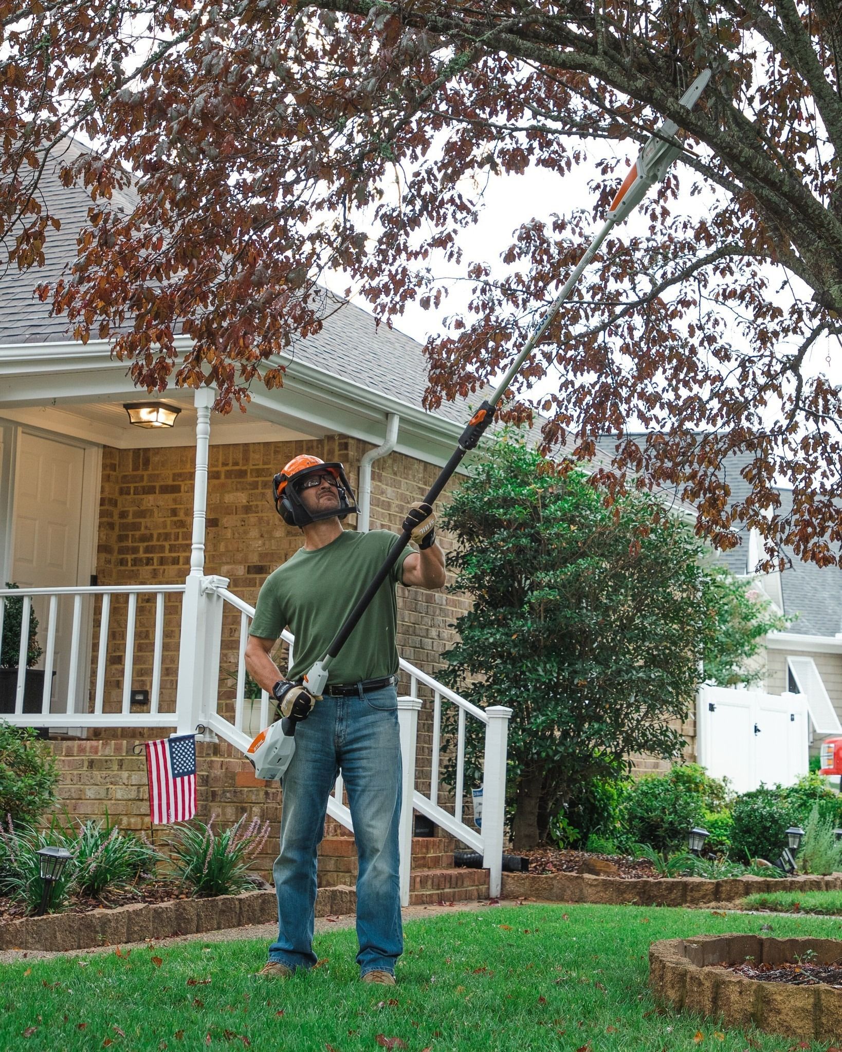Man using a pole saw to trim a tree in a residential yard. He wears a helmet and holds the saw overhead.