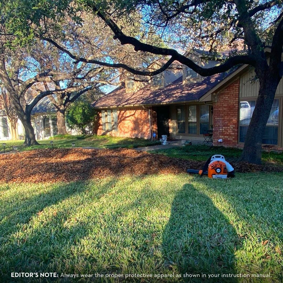 A person uses a leaf blower to clear leaves from a yard with a house in the background.