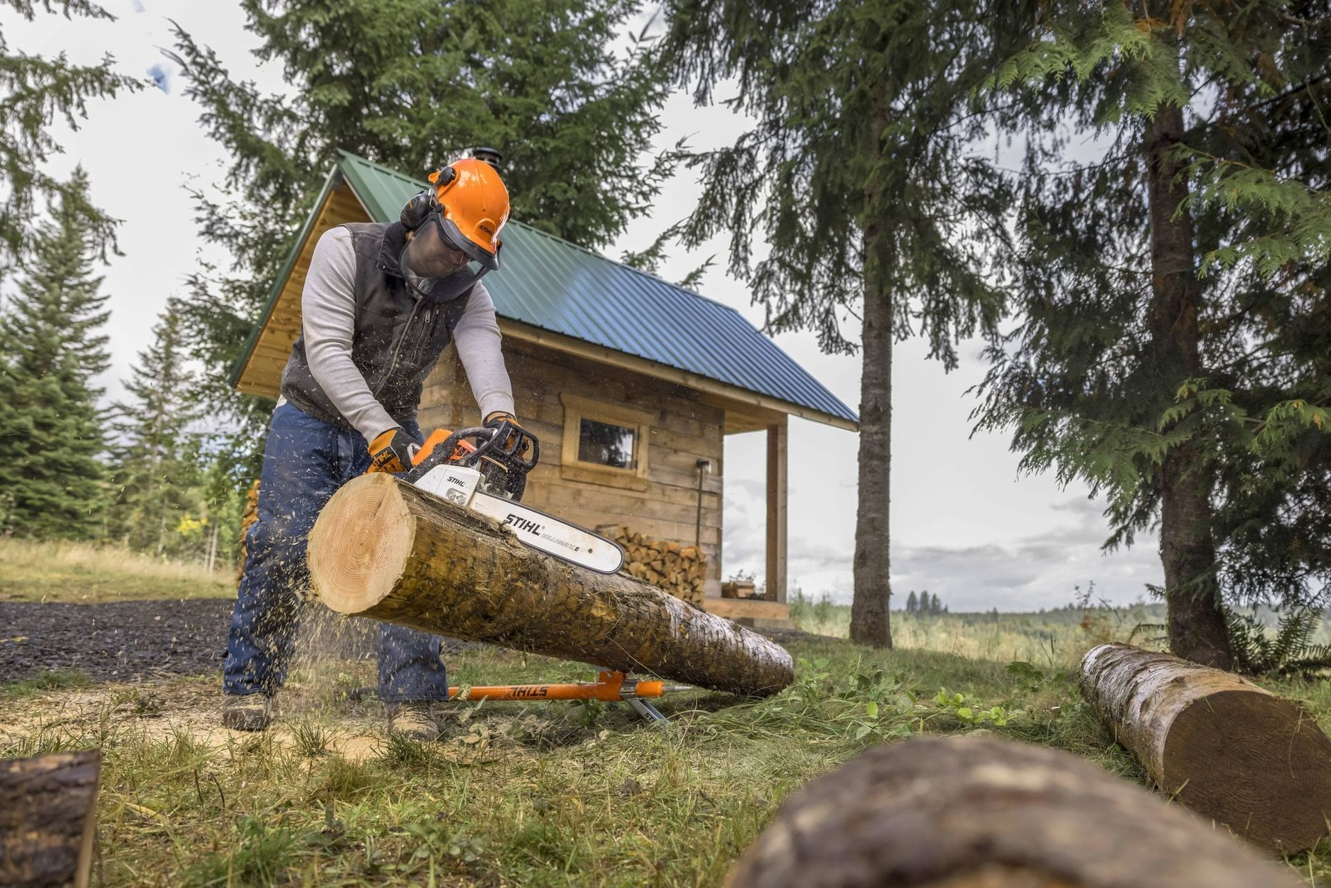 Person using a chainsaw to cut a log outdoors, wearing safety gear. Small cabin in the background.
