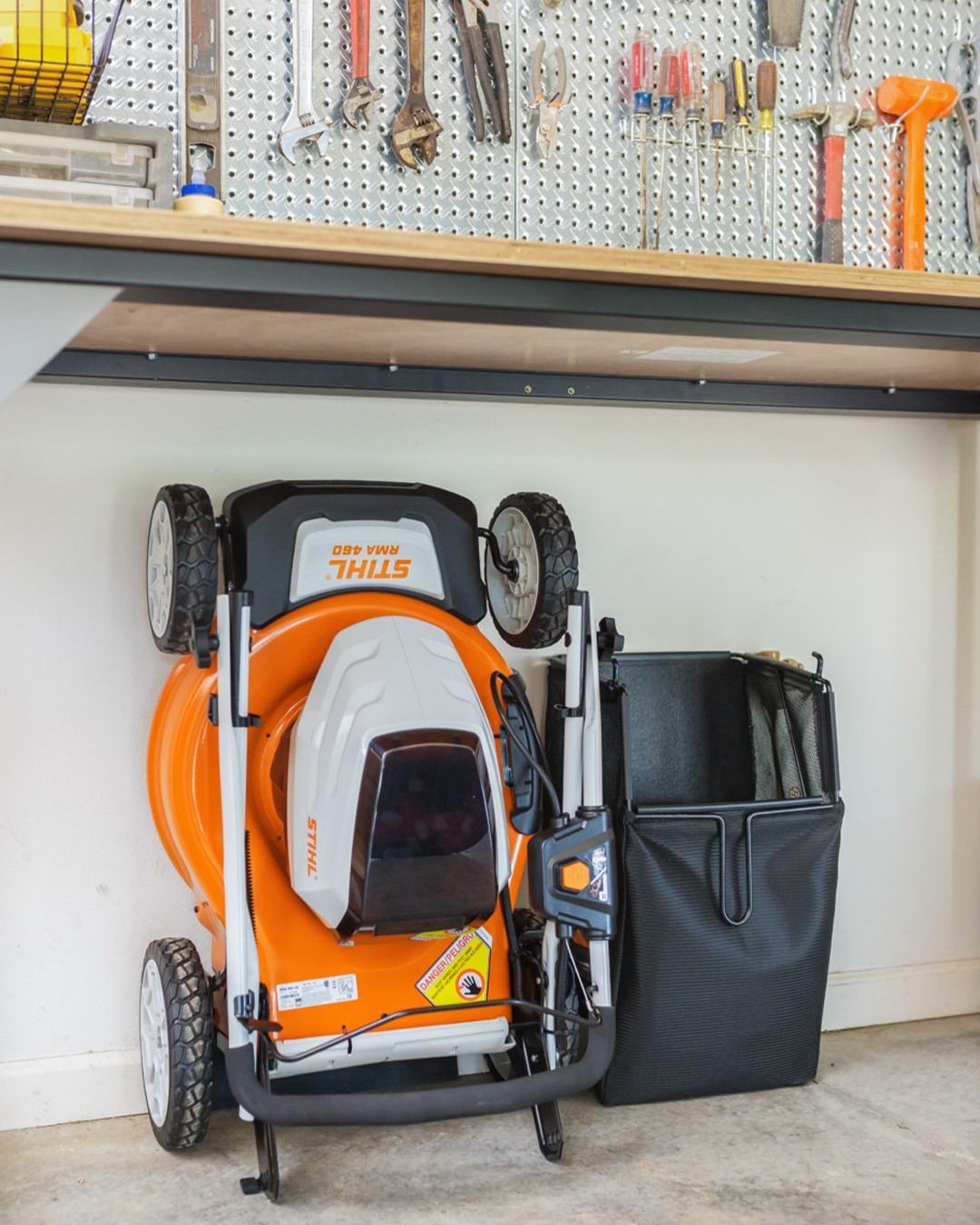 Orange and white lawnmower, folded, next to a black grass bag, under a workbench with tools.