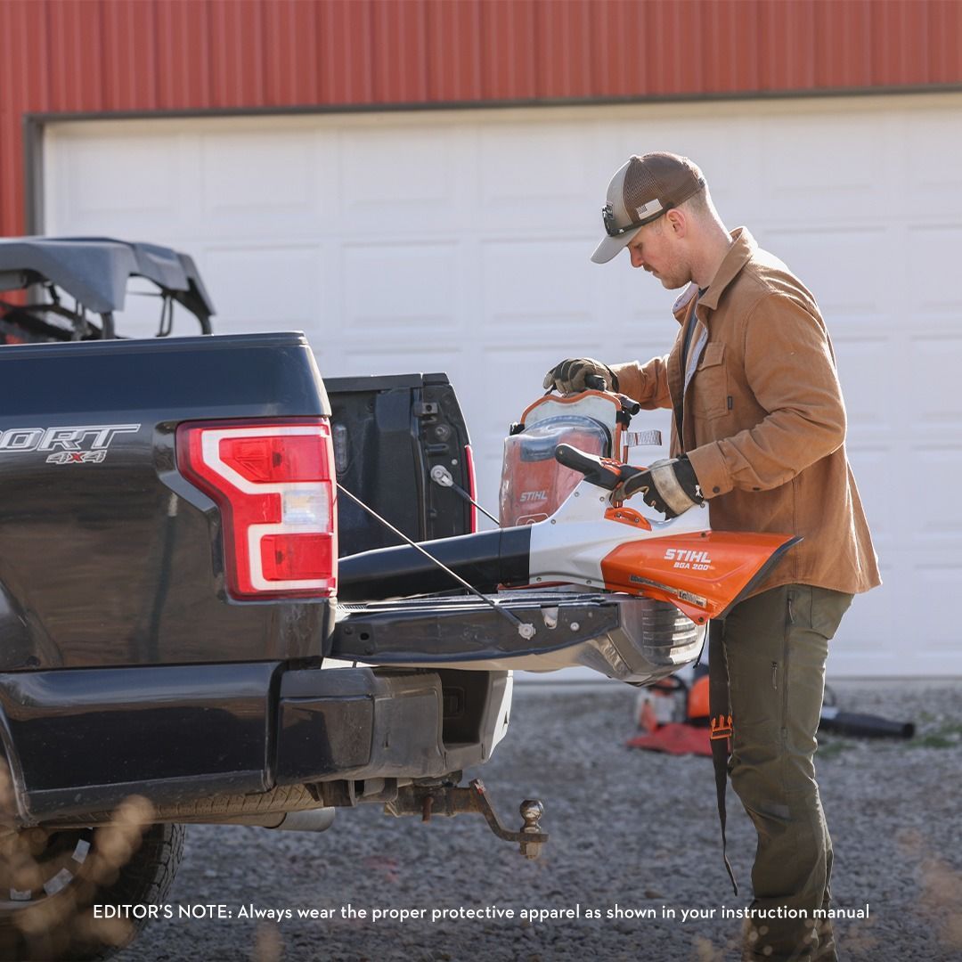 Man preparing a blower from a truck bed. He wears gloves, and a cap. Brown and orange equipment in setting.