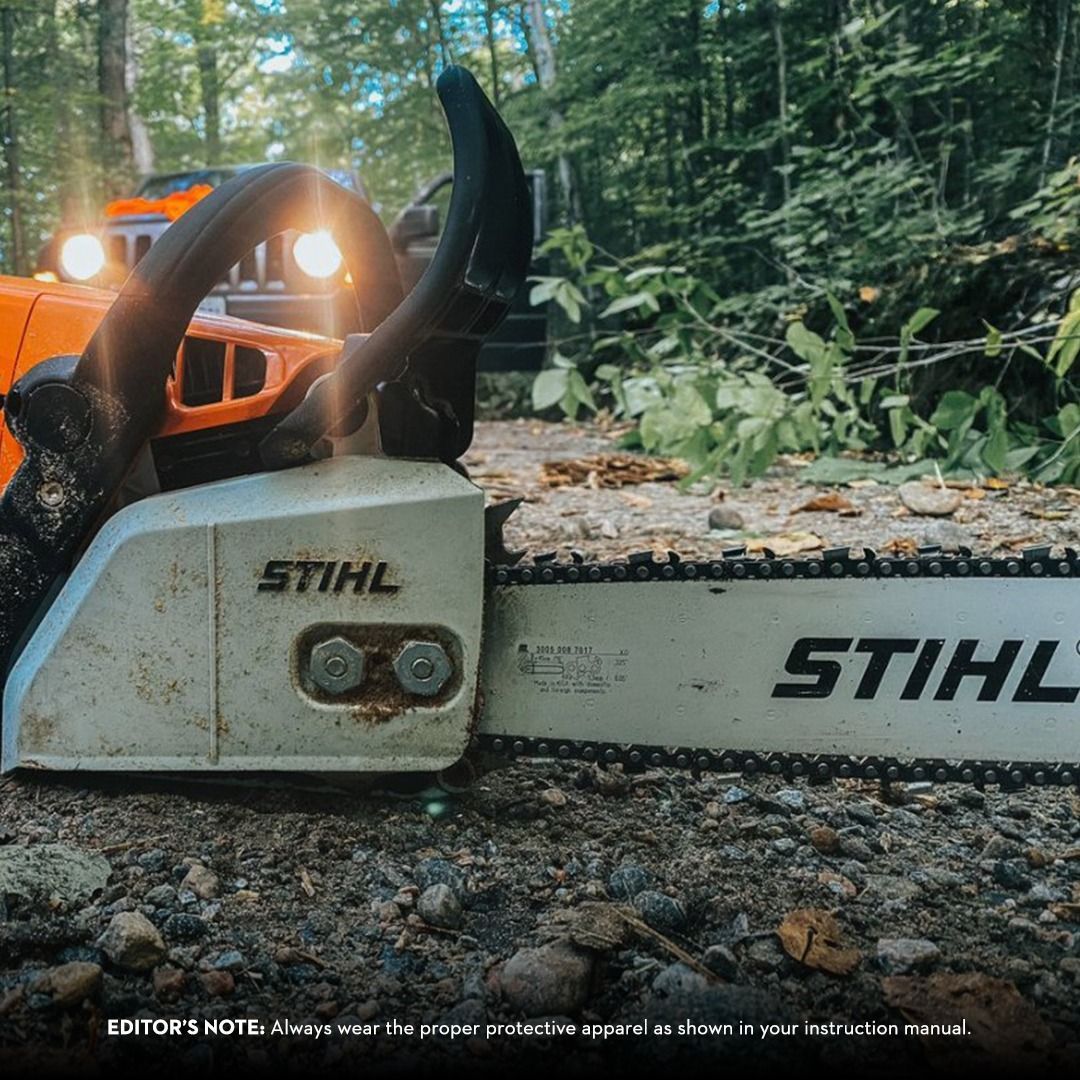 Stihl chainsaw on the ground with a Jeep in the background; outdoor setting, trees, and sunlight.