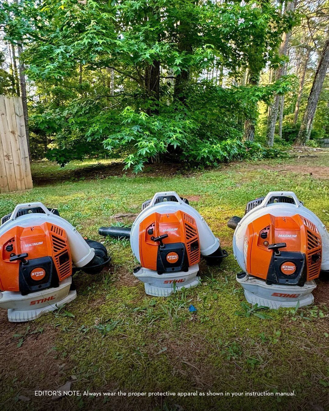 Three orange and white leaf blowers on green grass, trees in the background.