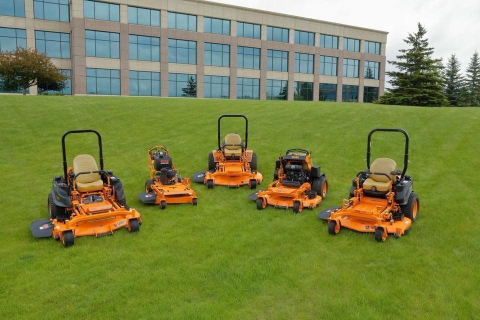 Five orange zero-turn mowers lined up on a green lawn in front of a building.
