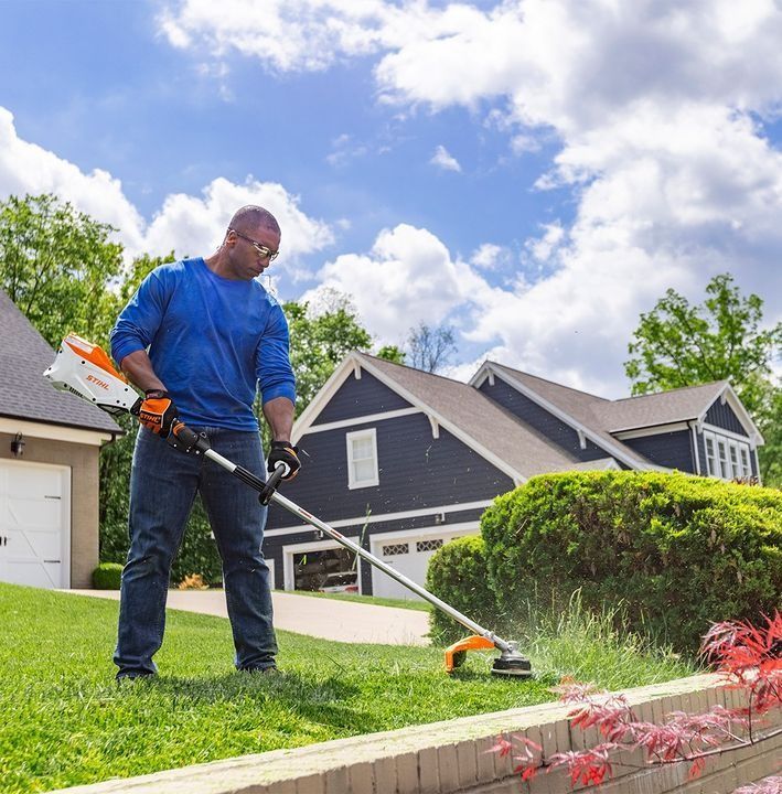 Man using a string trimmer on a lawn in front of a house. Sunny day with blue sky.