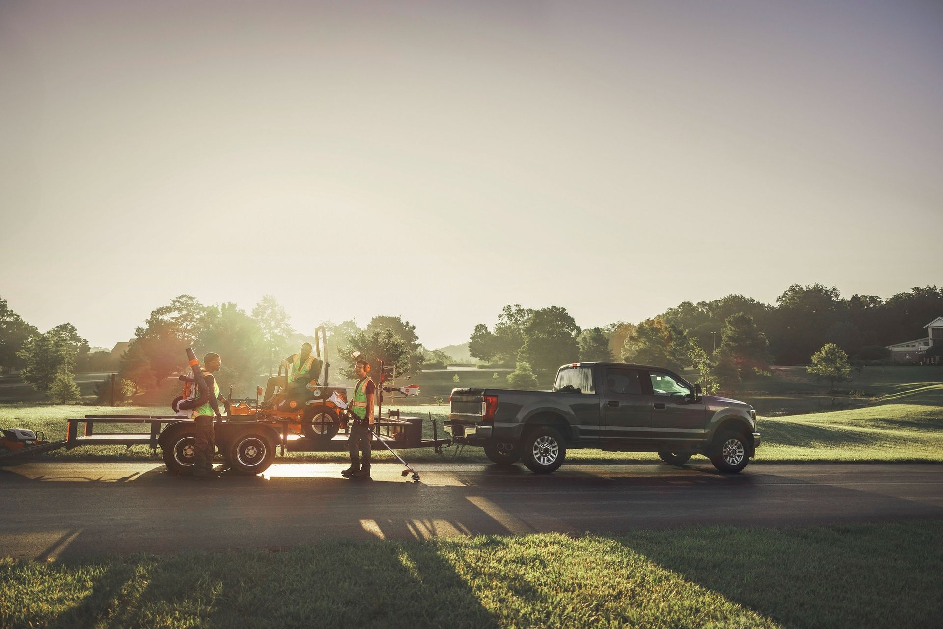 Truck towing a trailer with landscaping equipment on a road at dawn.
