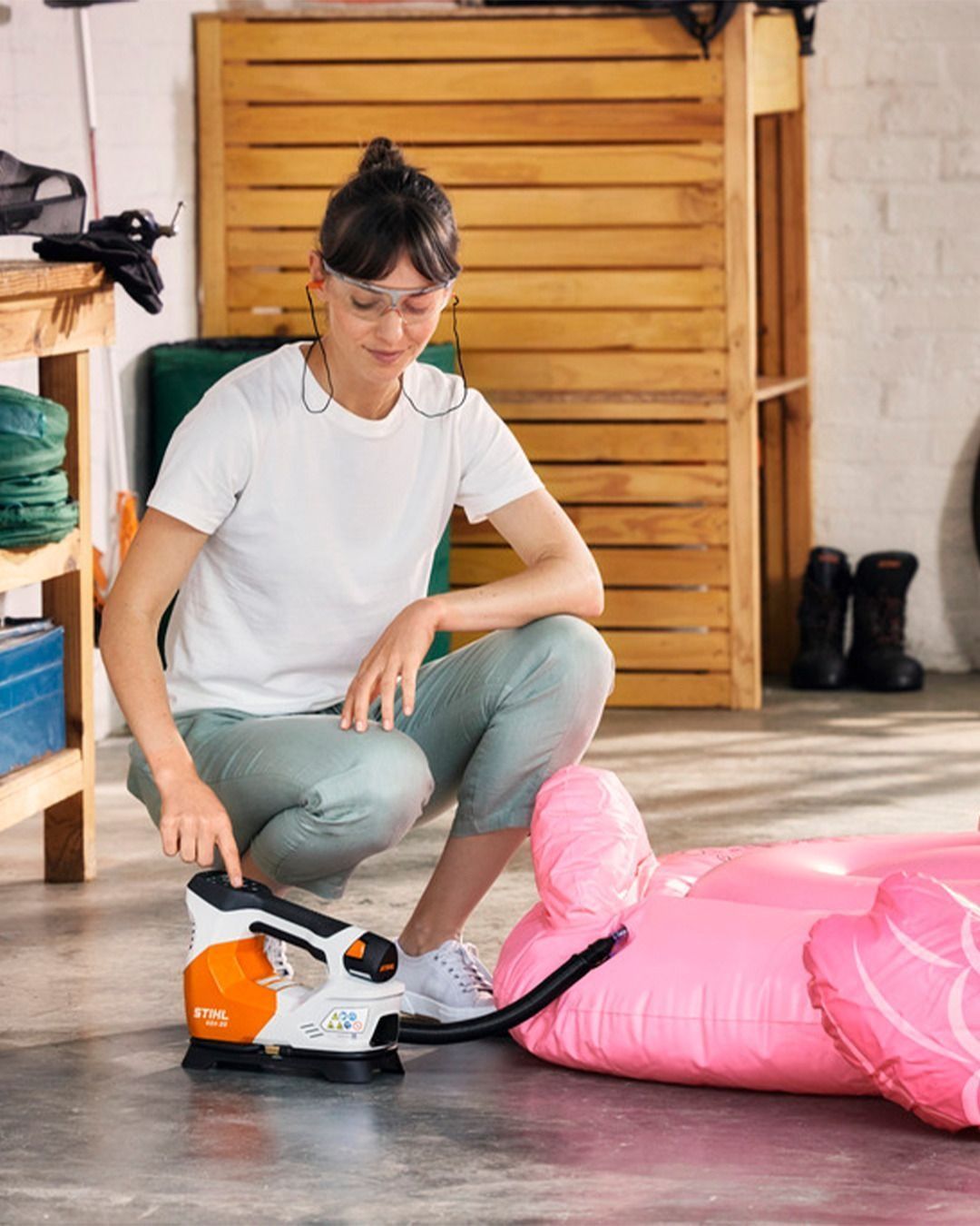 Woman inflating a pink flamingo float with an orange and white inflator in a garage.