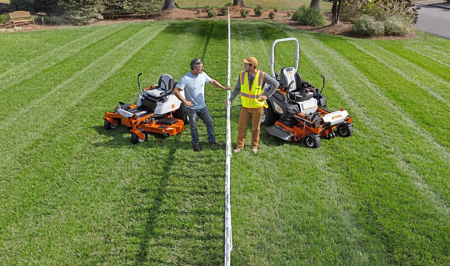 Two people stand between two zero-turn mowers on a striped lawn, holding a white pole.