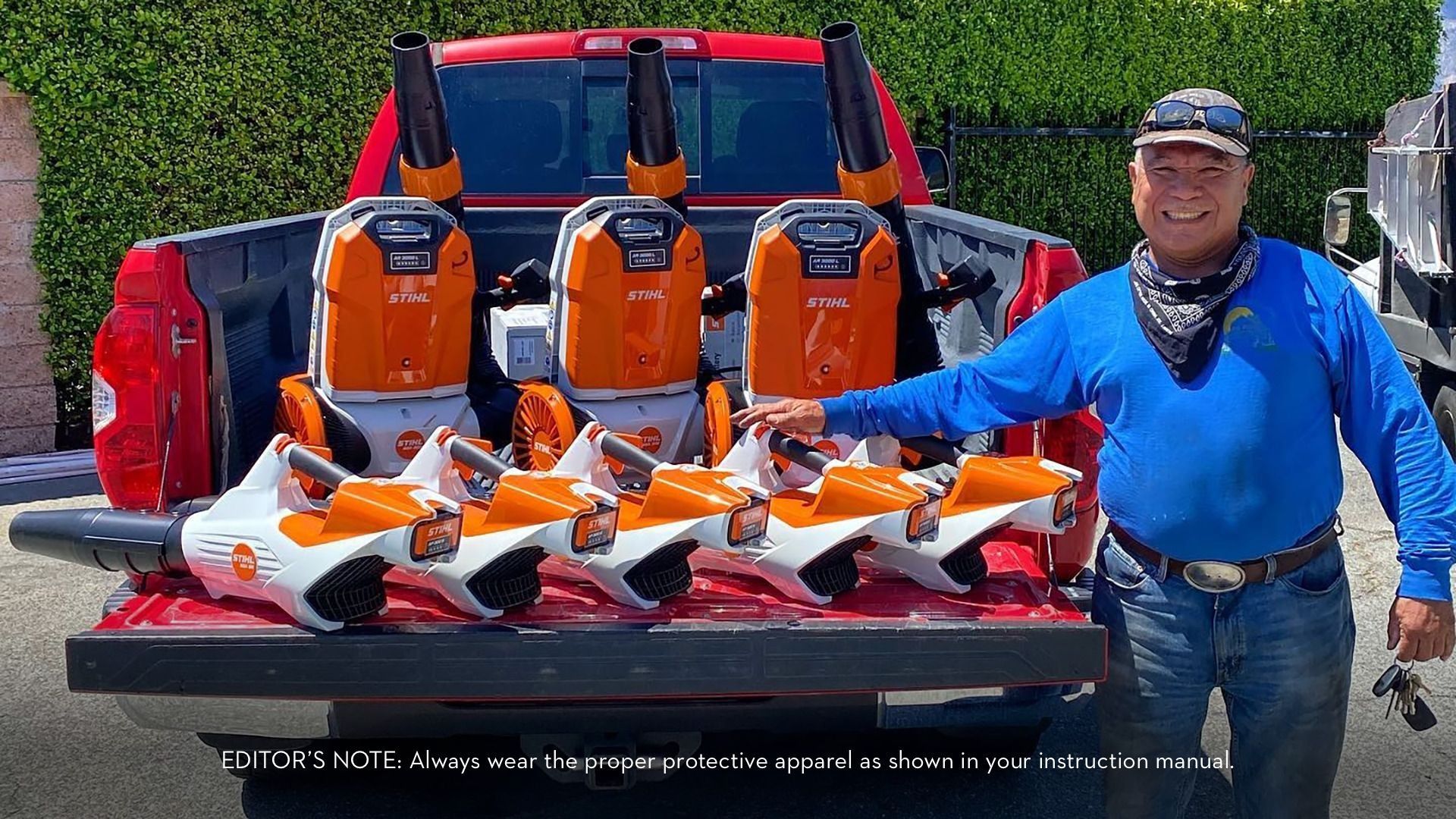 Man with leaf blowers in a truck bed. Orange and white equipment. Outdoors, sunny day.