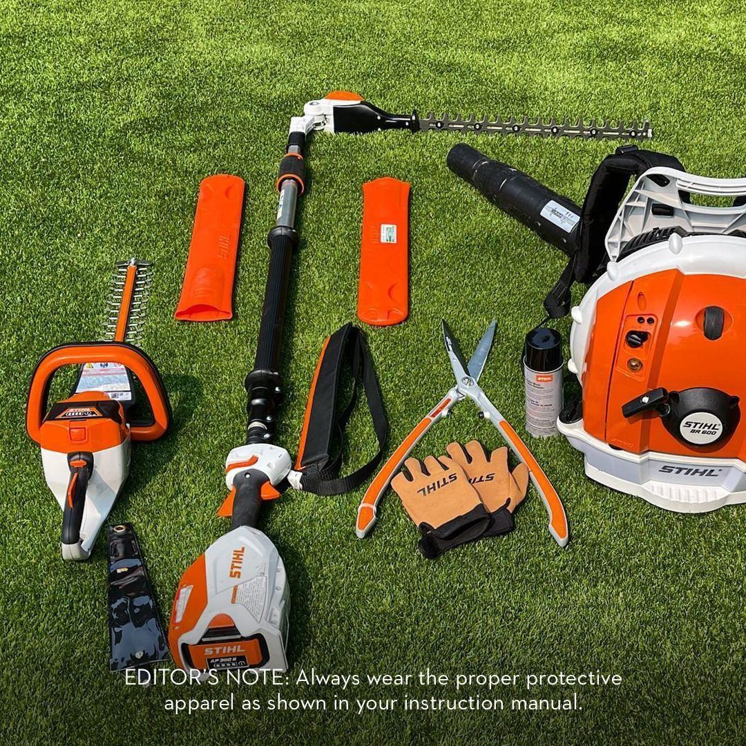Various orange and white Stihl yard tools laid on green grass. Includes a backpack blower, trimmer, and hand tools.