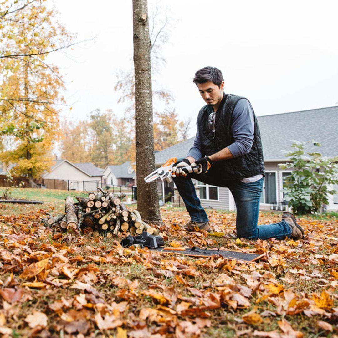 Man kneels in yard with a chainsaw, preparing to cut a tree amidst autumn leaves; houses in background.