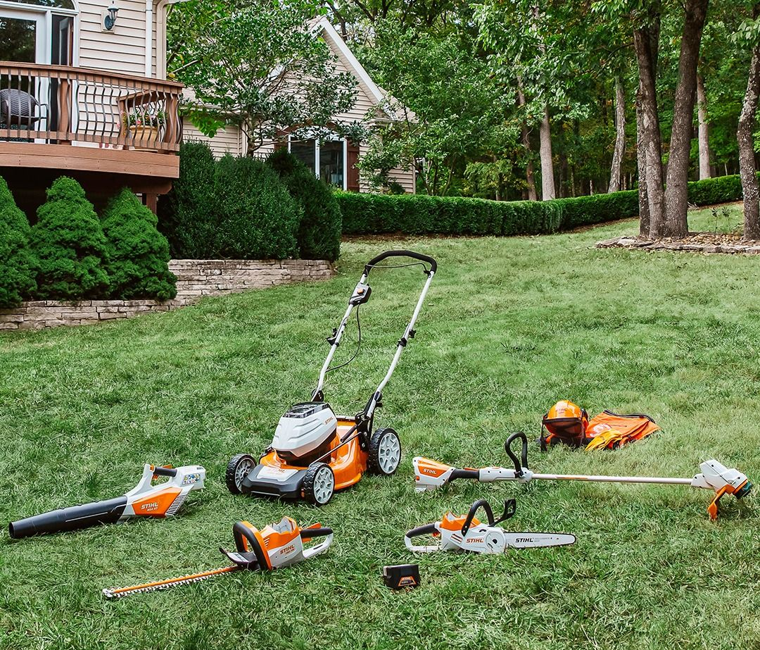 Assortment of STIHL yard tools on a lawn: mower, blower, trimmer, chainsaw, hedge trimmer, and battery.